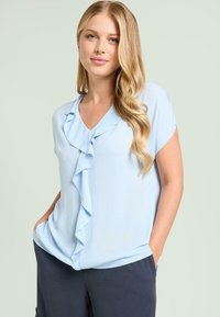 Lyseblå bluse med korte ærmer, V-hals og flæsedetalje foran. Glat stof, afslappet pasform, kombineret med mørke bukser.