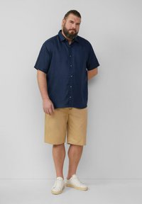 Marineblaue Kurzarmhemden mit Knöpfen, kombiniert mit beigen Shorts. Das Modell trägt weiße Turnschuhe und steht vor einem hellen Hintergrund.
