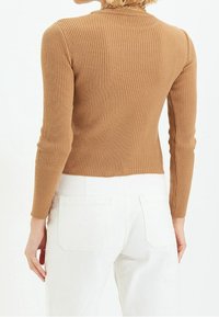 Haut à manches longues en maille côtelée de couleur marron clair, présentant une silhouette ajustée. Associé à un jean blanc, mettant en valeur la texture du tissu et la coupe.