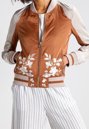 Chaqueta bomber marrón con mangas beige y bordado floral blanco en el delantero y los puños, combinada con pantalones blancos con rayas verticales grises.