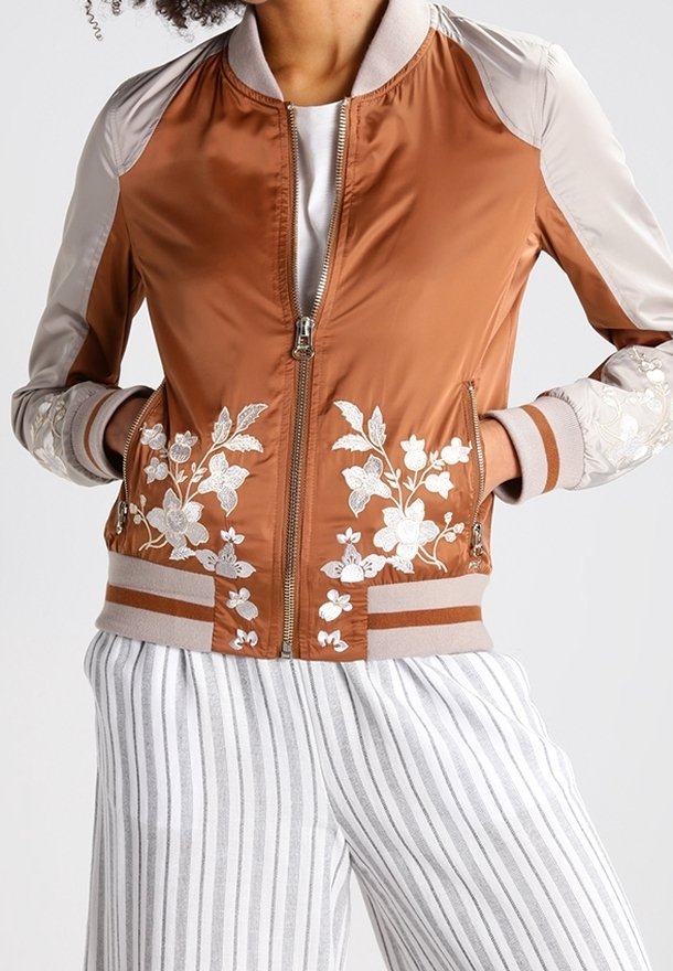 Veste bomber marron avec manches beiges et broderie florale blanche sur le devant et les poignets, portée avec un pantalon blanc à rayures verticales grises.