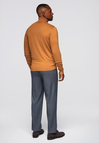 Brauner Rollkragenpullover, tailliert, kombiniert mit hellgrauen Hosen. Die Hosen sind gerade geschnitten und haben eine glatte Textur. Monte-braune Schuhe runden das Outfit ab.