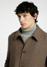 Cappotto a scacchi marrone con colletto a camicia e chiusura con bottone, indossato su un dolcevita grigio a coste. Tessuto morbido con una vestibilità su misura.