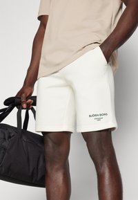 Weiße, weiche Stoffshorts mit lockerer Passform, featuring seitlichen Taschen und grünem bedrucktem Branding am linken Bein.