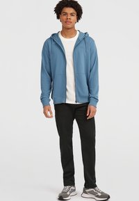 Felpa blu con zip e cappuccio regolabile, indossata sopra una maglietta bianca. Pantaloni neri e sneaker grigie completano l'outfit.