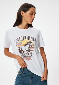 Biała bawełniana koszulka z grafiką brązowego konia, napisem "CALIFORNIA 1851" oraz paskami w kolorze pomarańczowym, żółtym i czerwonym, nawiązującymi do zachodu słońca. Krótkie rękawy.