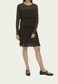 Schwarzes, gestuftes Kleid mit langen Ärmeln, aus strukturiertem Stoff mit dezentem Schimmer. Kombiniert mit schwarzen, flachen Schuhen mit dekorativen Details.