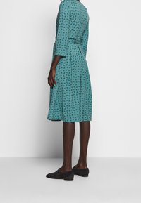 Robe turquoise à motifs avec manches trois-quarts, taille cintrée et ourlet à hauteur du genou. Portée avec des chaussures plates noires. Texture du tissu lisse.