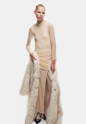 Robe longue, ajustée, nude avec un col haut, un buste drapé et une fente au niveau de la cuisse, associée à un manteau en fausse fourrure blanche et moelleux. Chaussures à talons visibles.