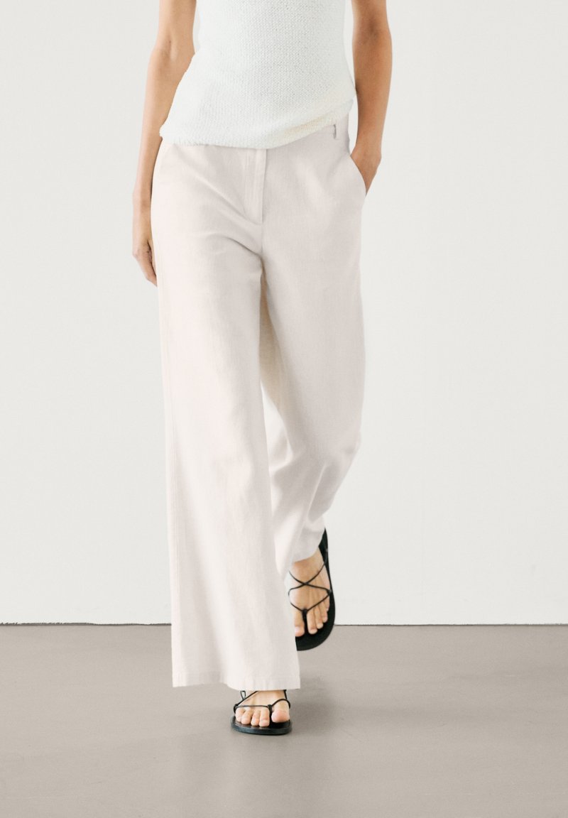 Femme portant un pantalon blanc ample et des sandales noires à brides, une main dans la poche, marchant sur un sol lisse sur fond blanc.