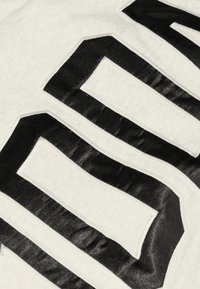 Weißer Stoff mit kräftigen, mattschwarzen Buchstaben. Der Text hat eine leicht erhabene Oberfläche, mit klaren Konturen und scharfen Kanten.
