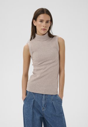 Frau trägt ärmelloses beigefarbenes Rollkragenoberteil und weite blaue Jeans, steht mit den Händen in den Taschen vor schlichtem Hintergrund.