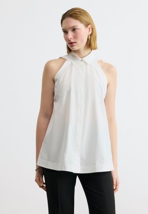 Γυναίκα που φοράει λευκό μπλουζάκι με άνετους ώμους και μαύρο παντελόνι, στέκεται μπροστά σε απλό λευκό φόντο.