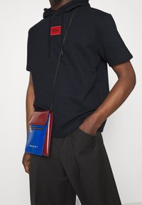 Mann trägt einen schwarzen kurzärmeligen Hoodie mit rotem Logo-Patch und einer kleinen quergetragenen Tasche aus blauem und bordeauxfarbenem Leder, Hand in der Tasche der dunklen Hose.