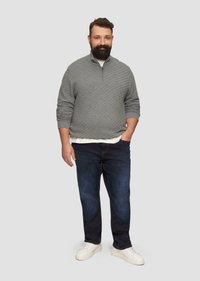 Szary sweter na zamek z kołnierzem, z wzorem w diamenty, biały t-shirt pod spodem, ciemnoniebieskie dżinsy i białe sneakersy. Model stoi na jasnym tle.