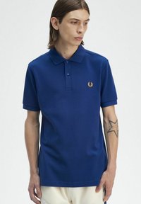Polo blu con colletto classico, tre bottoni e un piccolo logo ricamato in oro sul petto. Tessuto morbido e texturizzato.