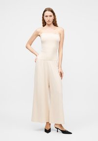 Νέα γυναίκα με κυματιστά μαλλιά, φορούντα ένα μπεζ ολόσωμο κορμάκι χωρίς ράντες και μαύρες μυτερές γόβες, στέκεται μπροστά σε ένα απλό λευκό φόντο.