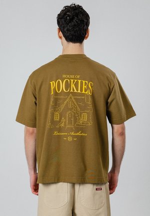 Mand med mørkt krøllet hår iført olivengrøn t-shirt med gul husillustration og teksten "House of Pockies Leisure Aesthetics" på ryggen.