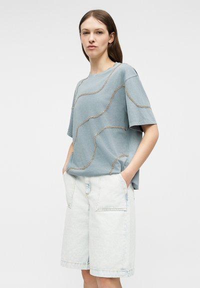 Γυναίκα με ανοιχτό μπλε φαρδιά μπλούζα με διακοσμητικές χάντρες και άνετο λευκό τζιν σορτς μέχρι τα γόνατα, με τα χέρια στις τσέπες.