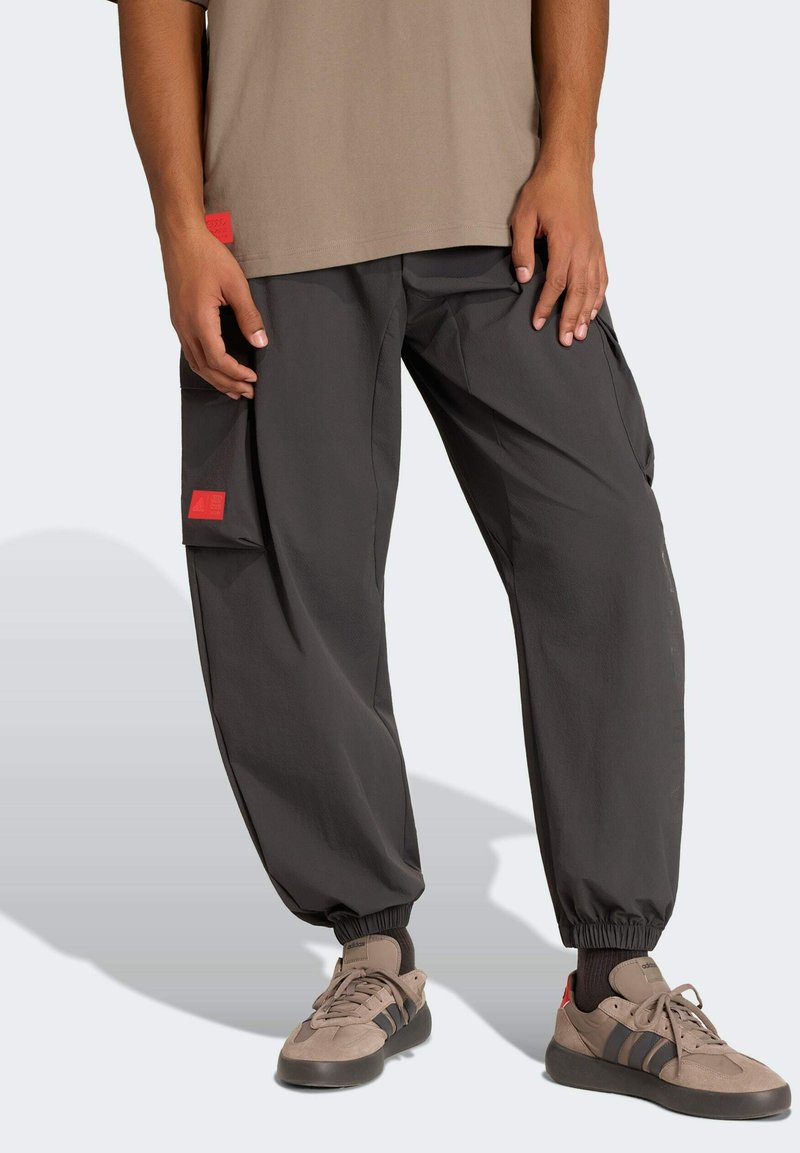 Person trägt schwarze Cargo-Joggerhosen mit elastischen Bündchen und beige Adidas-Sneaker mit schwarzen Streifen, steht vor einem schlichten Hintergrund.