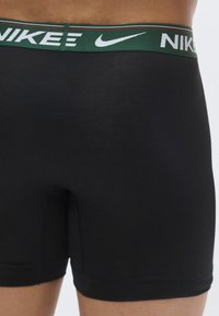 Boxeri negri cu talie elastică verde, având logo-ul alb „NIKE” și linii accentuate roșii în partea de sus. Textură netedă.