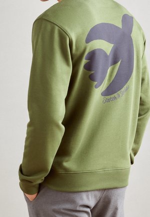 Mand iført olivengrøn sweatshirt med stort mørkt abstrakt fuglegrafik og lille kursiv tekst på ryggen, grå bukser, hænder i lommerne.