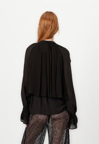 Zwarte blouse met een geplooide rug, lange mouwen en gerafelde manchetten, gemaakt van een lichtgewicht stof. Het ontwerp heeft subtiele textuur en lagen.