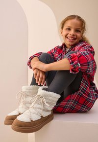 Bottes d'hiver blanches et duveteuses avec un design à lacets, semelles marron, et une texture douce. Portées avec des leggings foncés et une chemise à carreaux rouge.
