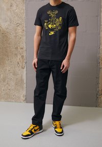 Černé bavlněné tričko s žlutým grafickým designem, v kombinaci s černými kalhotami a černobílými Nike teniskami. Jednoduchý, ležérní vzhled.