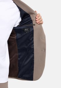 Beige blazer met marineblauwe satijnen voering, voorzien van een pocket met een geruite rand en een label met "JEFF" en maat "50."