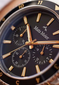 Montre chronographe avec un cadran noir, des index et aiguilles dorés, des sous-cadrans texturés et le logo "Mats Meier" au centre. Lunette en acier inoxydable argenté.