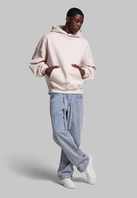 Overdimensioneret lys beige hoodie med en kængurulomme, parret med løse lyseblå jeans og hvide sneakers. Glat tekstur, afslappet stil.