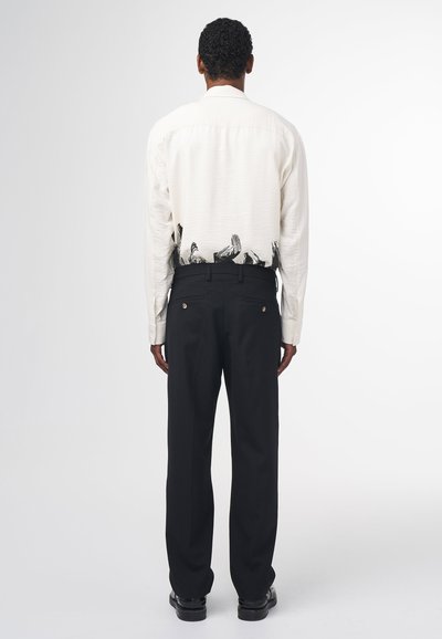 Weißes Langarmhemd mit einem schwarzen handgezeichneten Design am Saum, kombiniert mit schwarzen Hosen und schwarzen Schuhen. Glatte Textur, taillierte Passform.