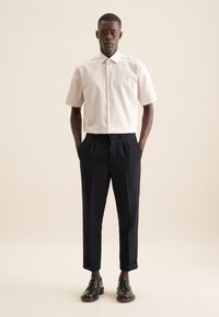 Camicia a maniche corte a righe in rosa pallido e bianco, abbinata a pantaloni scuri a vita alta in blu navy. La modella indossa scarpe in pelle nera.