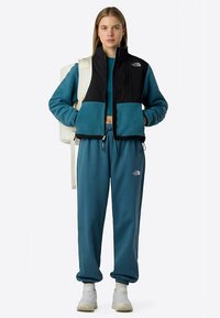 Fleecevest in zwart en teal, gecombineerd met bijpassende tealbroek. Witte sneakers en een lichte rugzak. Textuurdetails van de stof zichtbaar.