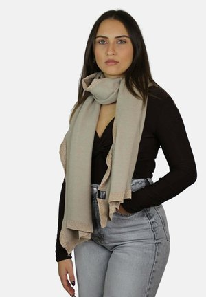 Jeune femme aux cheveux longs portant une écharpe beige sur un haut noir et un jean gris clair taille haute, debout devant un fond blanc uni.