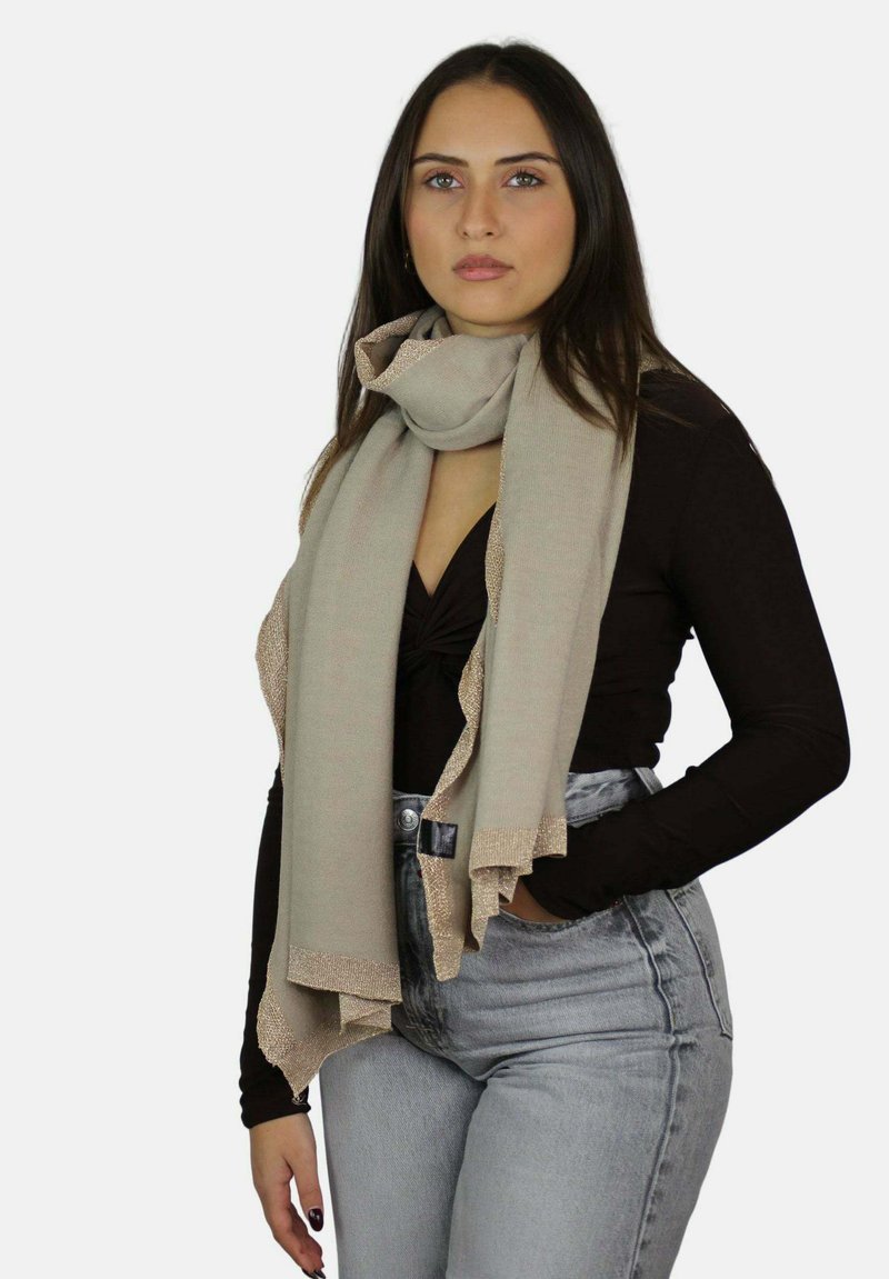 Młoda kobieta z długimi włosami ma na sobie beżowy szalik, czarną bluzkę i jasnoszare wysokowalne spodnie, stoi na tle jednolitego białego tła.