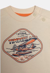 Beige sweatshirt med en grafik av ett sjöflygplan och texten "TIMBERLAND" i orange och marinblått. Ribbad hals med tryckknappar.