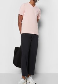 Camisa de manga corta en tono rosa claro con escote en V y un logo en el pecho, combinada con pantalones de pierna ancha negros y zapatillas blancas, llevando una bolsa tote negra.