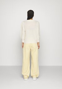 Biały sweter z dzianiny z bufkami, w połączeniu z szerokimi spodniami z jasnej żółtej cordurowej tkaniny, na jednolitym tle.