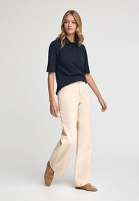 Granatowa koszulka polo z krótkimi rękawami i trzema guzikami, sparowana z kremowymi spodniami o szerokich nogawkach i jasnobrązowymi mokasynami. Obserwowane gładkie tekstury.