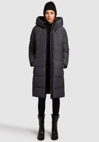 Dlhý tmavosivý prešívaný kabát s kapucňou, prešívaný dizajn, zapínanie na zips a predné vrecká. Nosený cez čierny outfit a doplnený čiernymi topánkami.