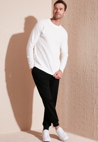Weißes Langarmhemd mit einem strukturierten Muster, kombiniert mit schwarzen Jogginghosen und weißen Sneakers, vor einer hellen strukturierten Wand stehend.