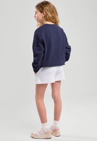 Sweat à capuche bleu marine à coupe décontractée, associé à un short gris clair. Porte des baskets New Balance roses avec des chaussettes blanches, mettant en valeur un style décontracté.