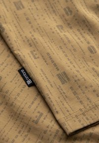 Tissu marron avec un motif de texte vertical subtil et une étiquette noire marquée « onlina » cousue près d’un bord plié.