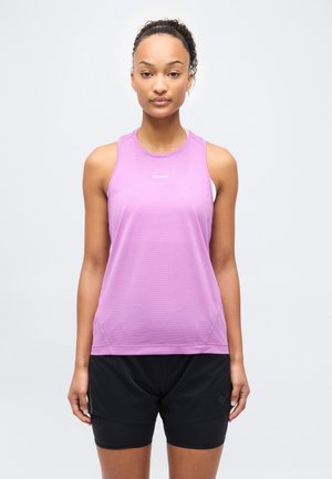 Jonge vrouw met krullend haar in een staart, gekleed in een paarse sporttop zonder mouwen en zwarte korte broek, staand tegen een effen witte achtergrond.