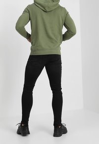 Olivengrøn hættetrøje med ribbet kant, kombineret med tætsiddende sorte jeans og sorte sportssko, set bagfra.