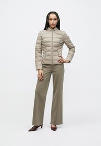 Bėgių smėlio spalvos dygsniuota striukė su aukštu apykakliu, užtrauktuku ir šoninėmis kišenėmis, derinama su alyvinių žalių platėjančių kelnių ir tamsiai raudonų aukštakulnių.