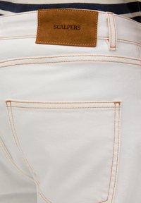 Jeans de mezclilla blanca con costuras en naranja claro y una etiqueta de cuero marrón con la marca "SCALPERS" en la parte trasera de la cintura. Dos bolsillos traseros visibles.