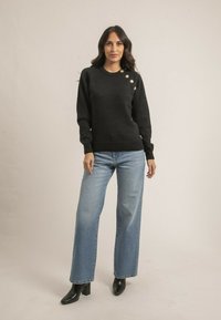 Pull en maille noir avec un ourlet côtelé et des manches longues, présentant des boutons dorés sur une épaule. Associé à un jean taille haute bleu clair.